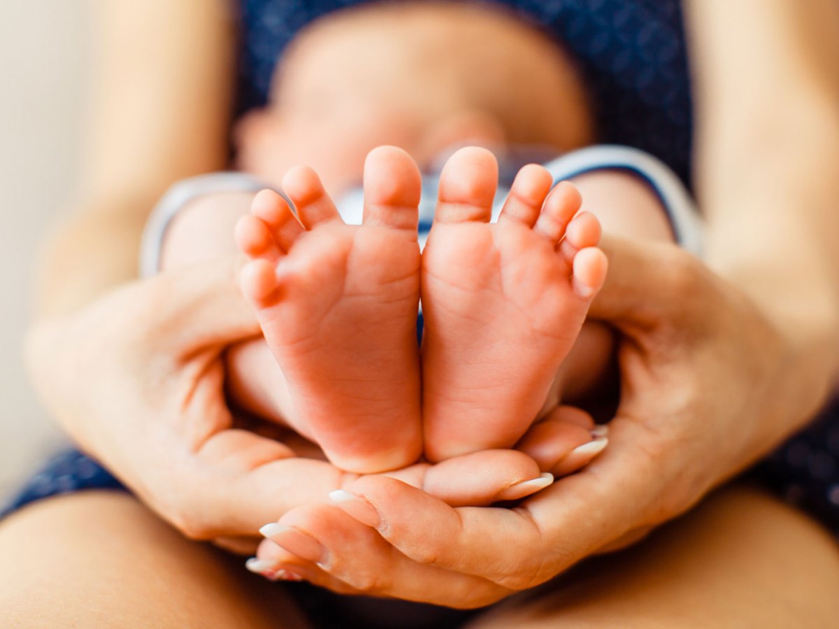 Nahaufnahme von Babyf&uuml;&szlig;en, die von den H&auml;nden der Mutter gehalten werden. Im Hintergrund erkennt man verschwommen, dass ein Baby auf dem Scho&szlig; seiner Mutter liegt.