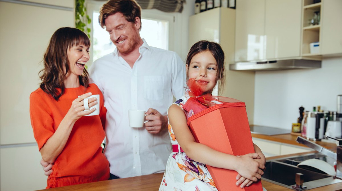 Familie in der Küche. Die Tochter hält ihre Schultüte, die Eltern stehen dahinter, lachen und halten eine Tasse in der Hand.