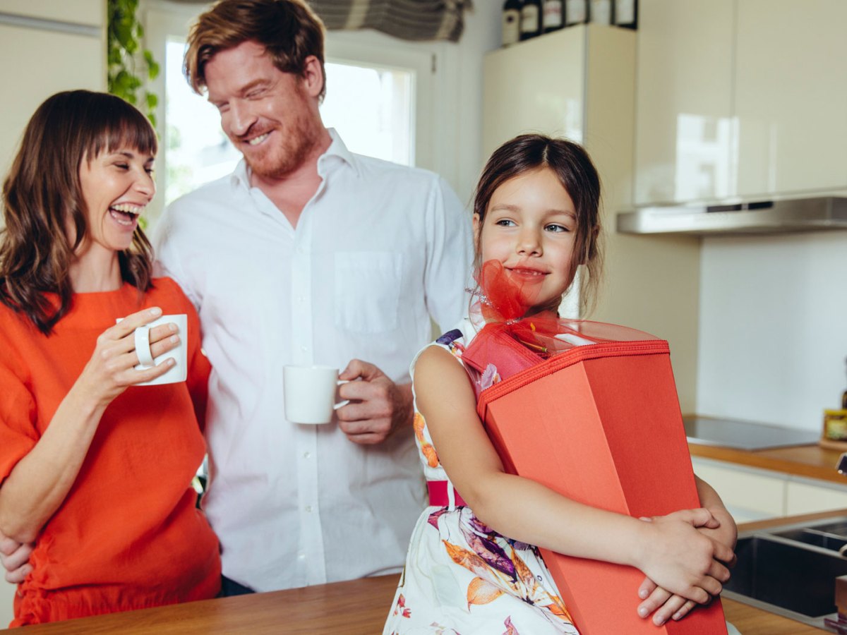 Familie in der Küche. Die Tochter hält ihre Schultüte, die Eltern stehen dahinter, lachen und halten eine Tasse in der Hand.