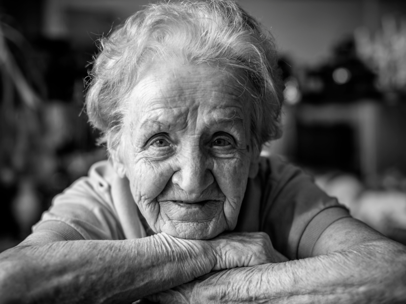 Eine alte Frau, die ihren Kopf auf den Armen abstützt und in die Kamera schaut in Schwarz weiß.
