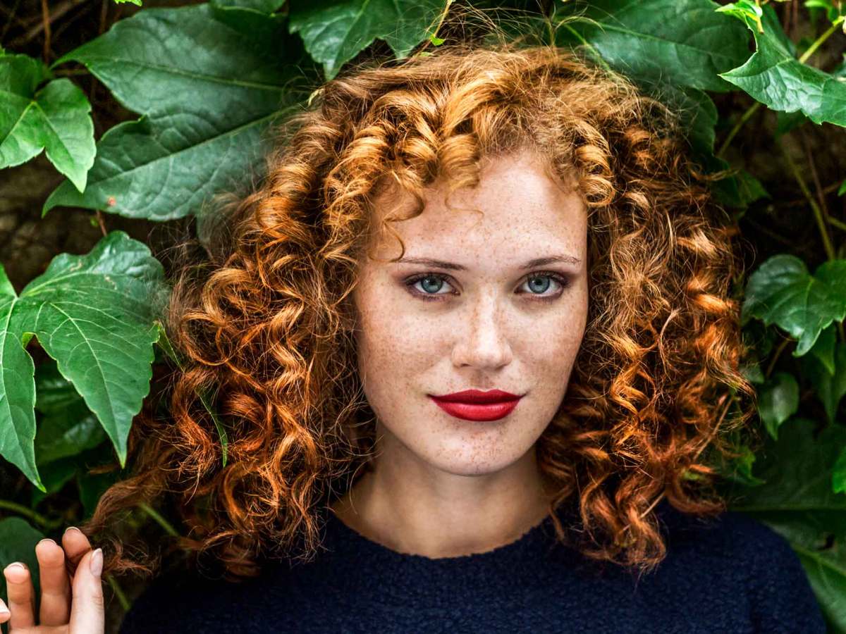 Junge Frau mit roten Locken, Sommersprossen und leuchtend roten Lippen blickt selbstbewusst vor einer grünen Blätterwand in die Kamera.