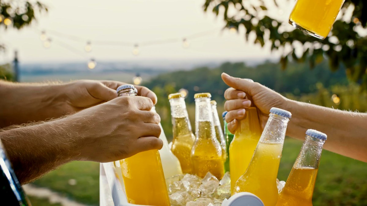 Mehrere Hände greifen nach gekühlten Flaschen mit gelbem Getränk aus einer mit Eis gefüllten Box im Freien, im Hintergrund Lichterketten und unscharfe Landschaft.