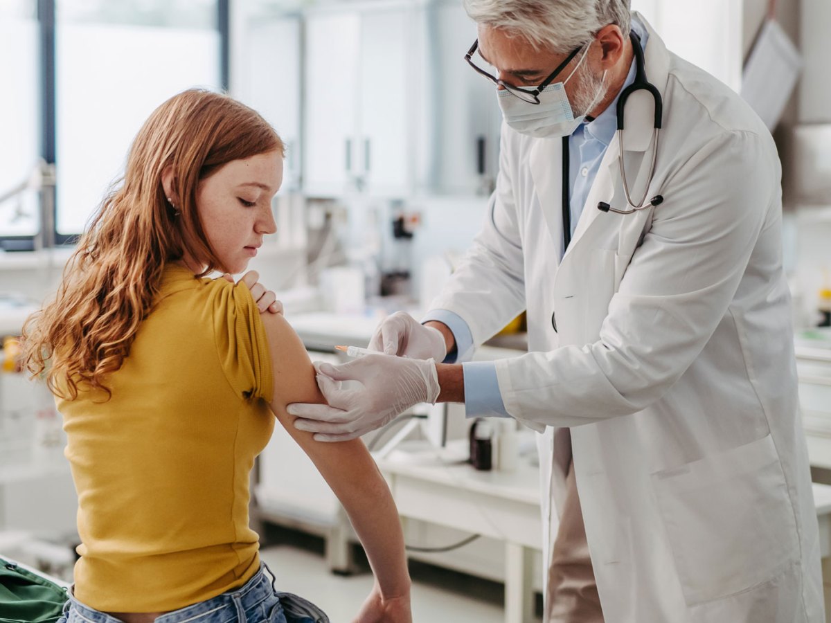 M&auml;dchen, ca. 13 Jahre, sitzt in der Arztpraxis und erh&auml;lt eine Spritze von einem &auml;lteren, m&auml;nnlichen Arzt.