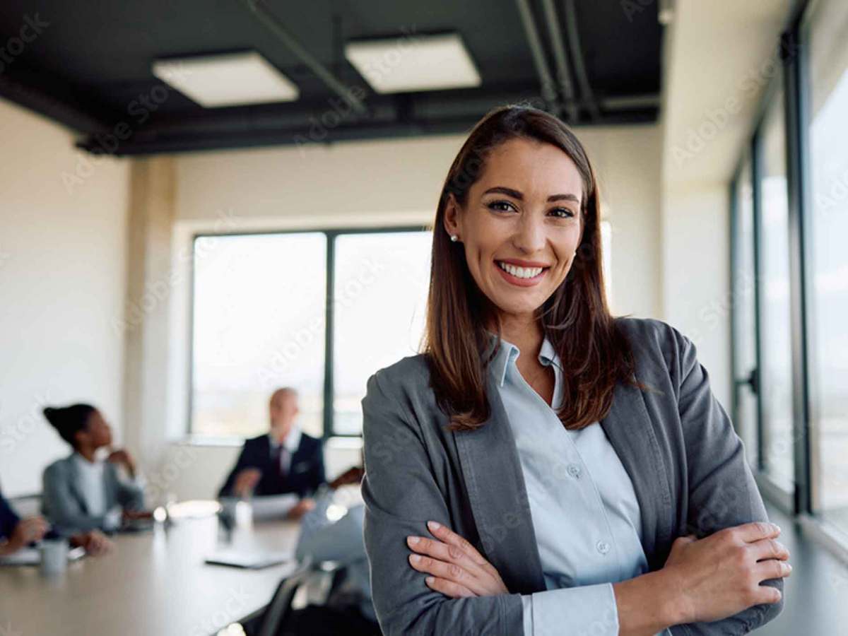 Braunhaarige Frau mit Blazer und verschr&auml;nkten Armen steht im B&uuml;ro und l&auml;chelt.