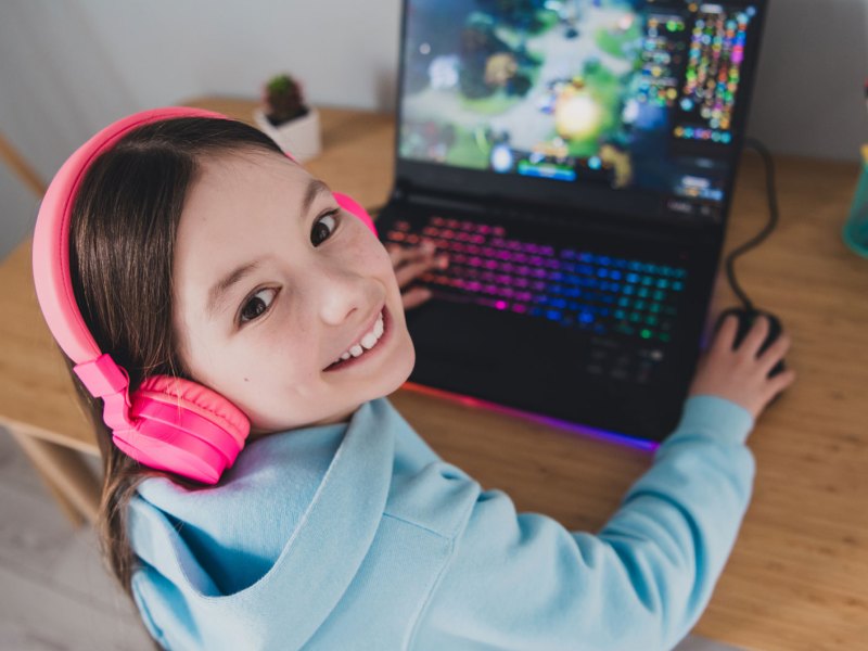 Mädchen, ca. 10 Jahre alt, sitzt mit Kopfhörern am Laptop und spielt darauf.