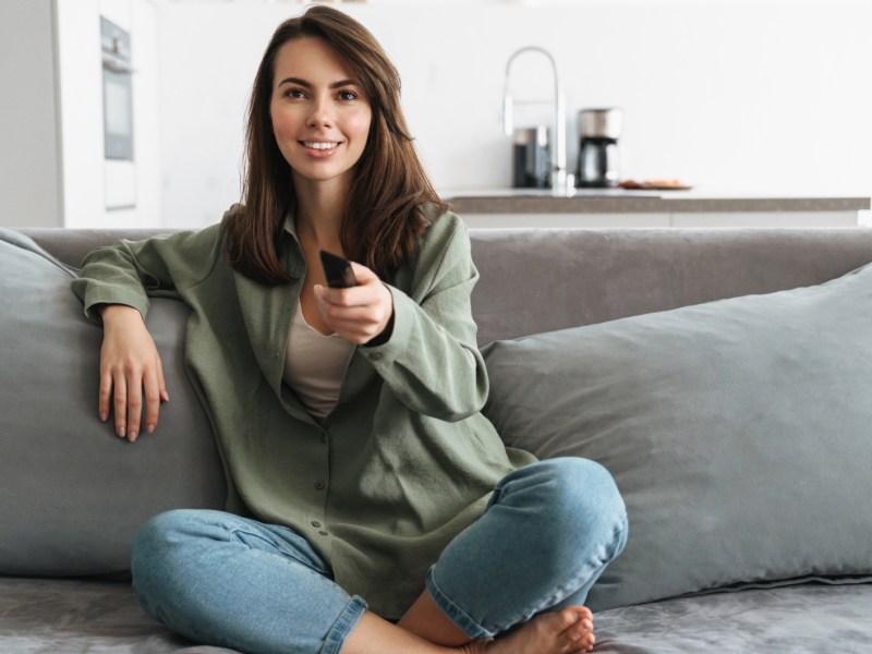 Brünette Frau sitzt auf grauem Sofa, die lächelt und hat Fernbedienung auf Kamera gerichtet.