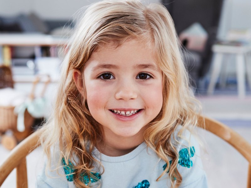 Porträt eines kleinen Mädchens, ca. 4 Jahre alt, das schmunzelnd an einem Tisch sitzt.