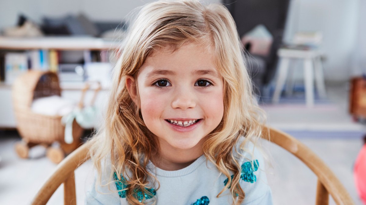 Porträt eines kleinen Mädchens, ca. 4 Jahre alt, das schmunzelnd an einem Tisch sitzt.