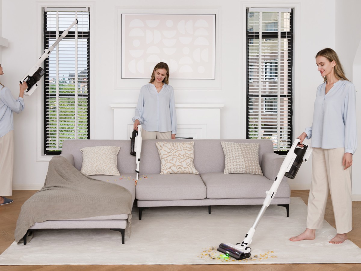 Eine Frau in hellblauer Bluse und beige Hose staubsaugt gleichzeitig den Teppich, das Sofa und die Jalousien in einem lichtdurchfluteten Raum.