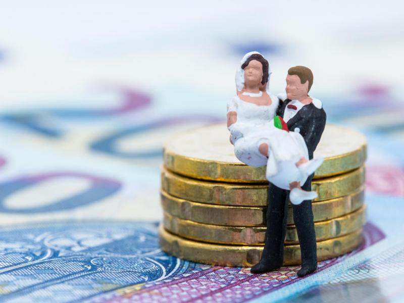 Kleine Brautpaarfiguren stehen auf Geldscheinen und neben einem Münzturm.