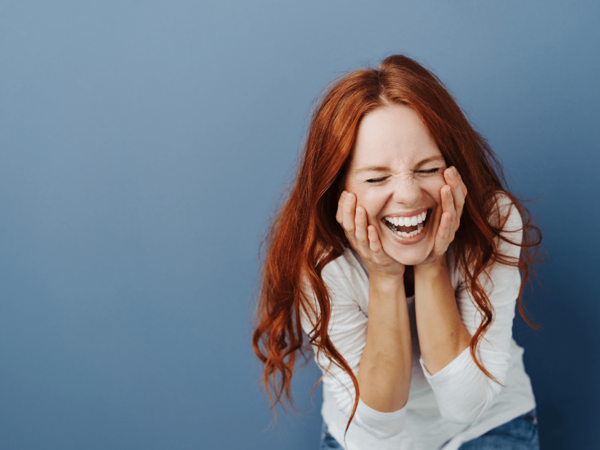 Frau hockt vor einem blauen Hintergrund mit roten Haaren und lacht