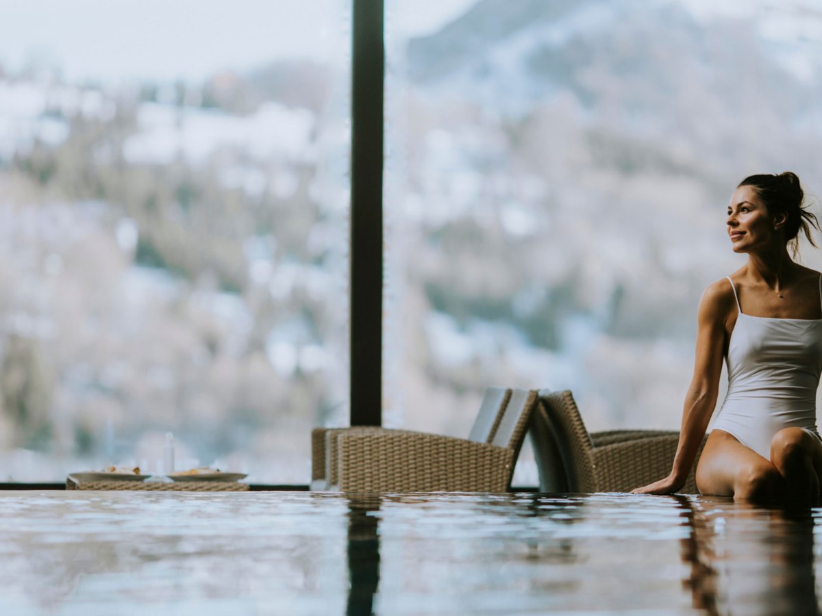 Frau sitzt auf Beckenrand eines Schwimmbeckens, in einem Wellnesshotel in den verschneiten Bergen.