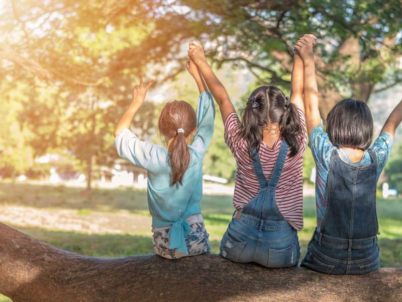 Drei Mädchen sitzen auf einem dicken Ast eines Baumes, haben ihre Hände in die Höhe gestreckt, halten einander fest und zeigen das Peace-Symbol.