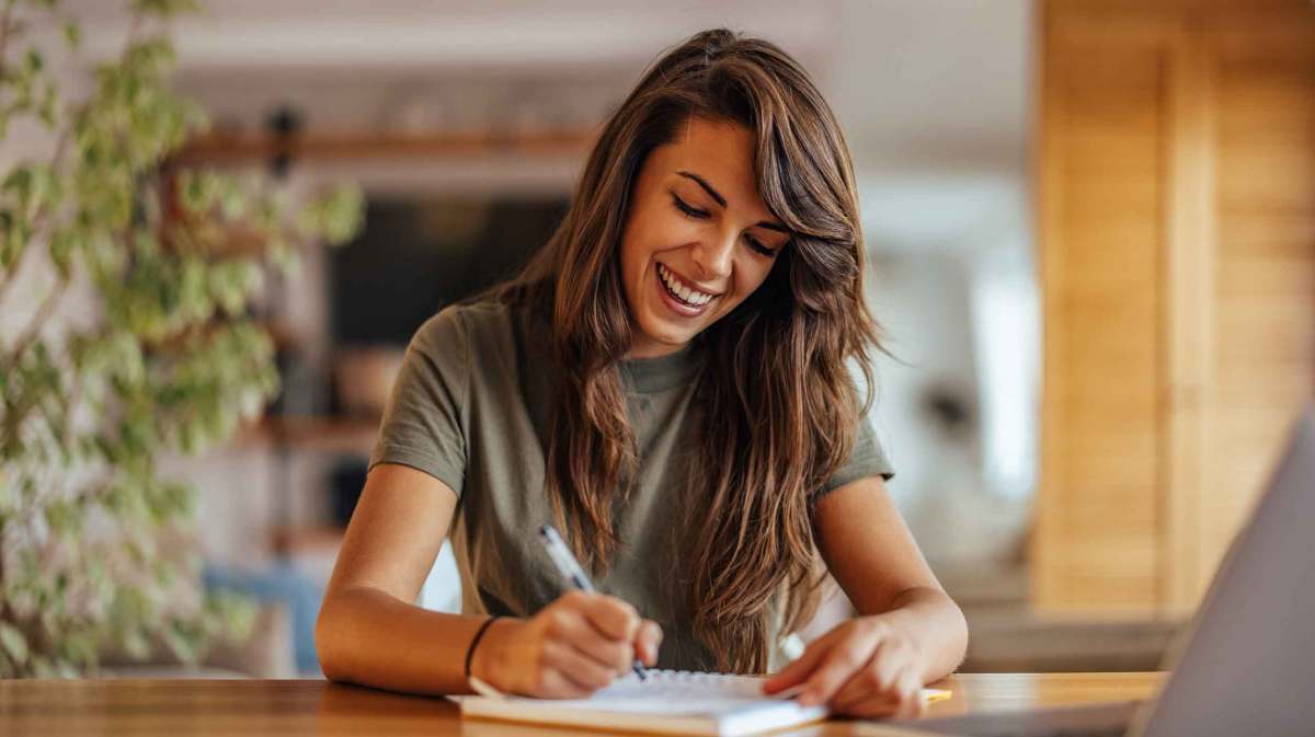 Frau sitzt an einem Schreibtisch und schreibt etwas auf und lacht dabei.