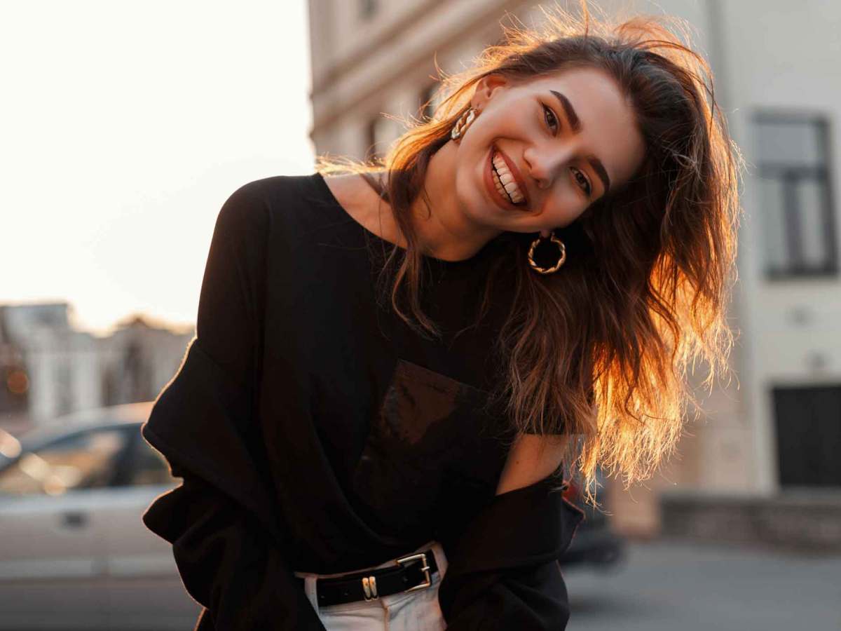 Junge Frau mit vollen Haaren steht drau&szlig;en und neigt den Kopf zur Seite und lacht.