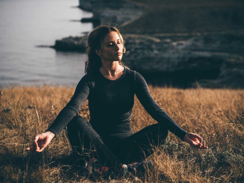 Junge Frau in schwarzem Hemd übt Yoga-Asanas auf hoher Felsenküste über dem Meer bei hellem Sonnenlicht und leichtem Wind in ruhigem Morgen