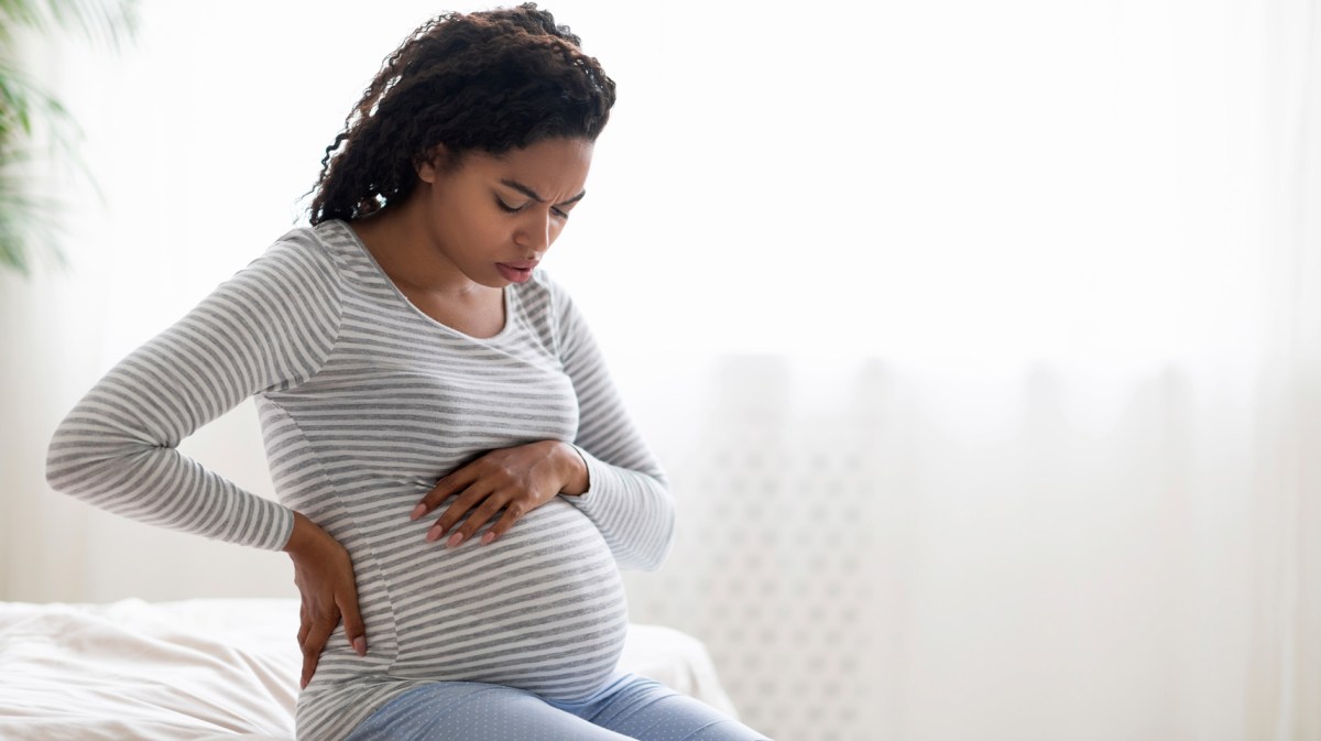 Schwangere Frau sitzt mit Wehen auf dem Bett zu Hause und hat eine Hand jeweils auf Bauch und Rücken gelegt.