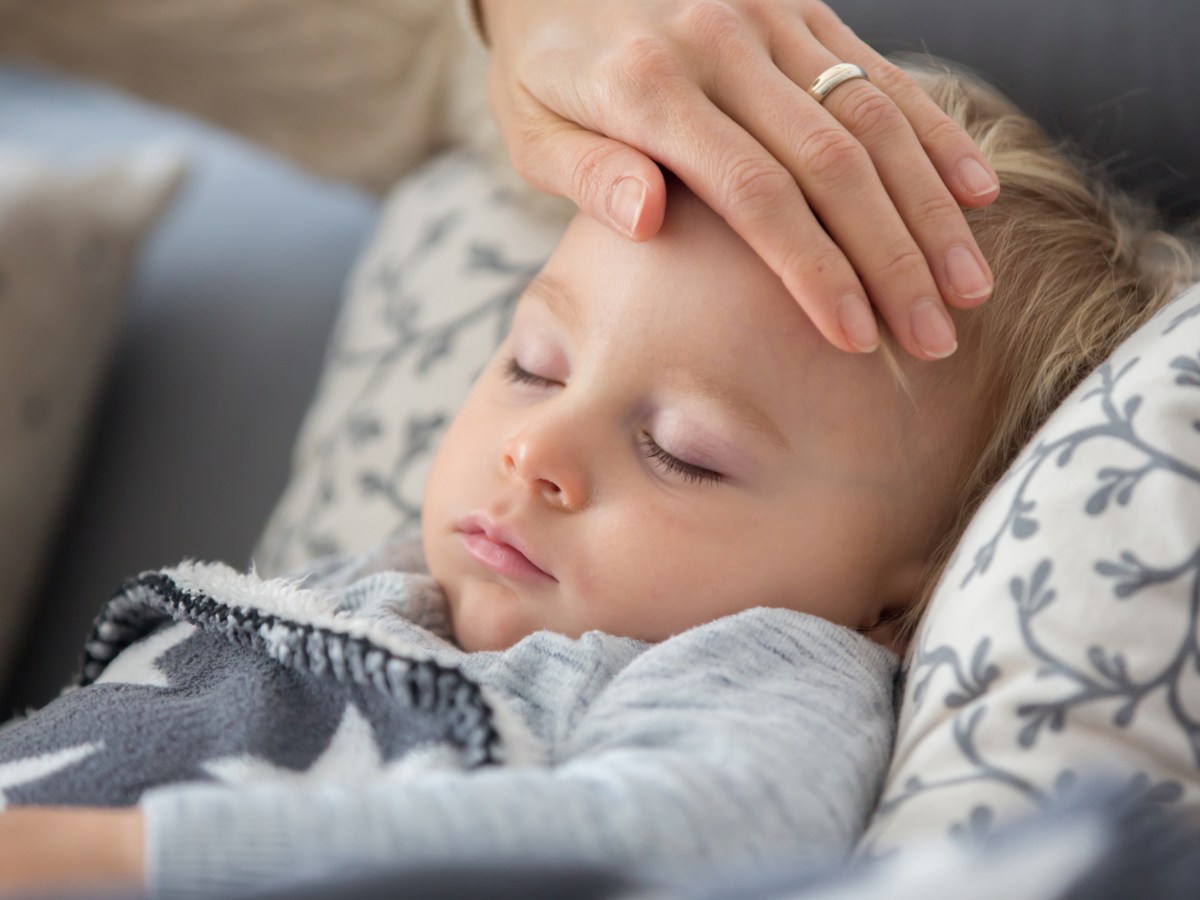 Krankes KInd liegt auf dem Sofa. Die Hand der Mutter fühlt den Kopf.