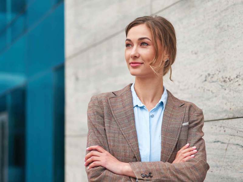 Frau mit Blazer steht mit verschränkten Armen vor einem Bürogebäude.