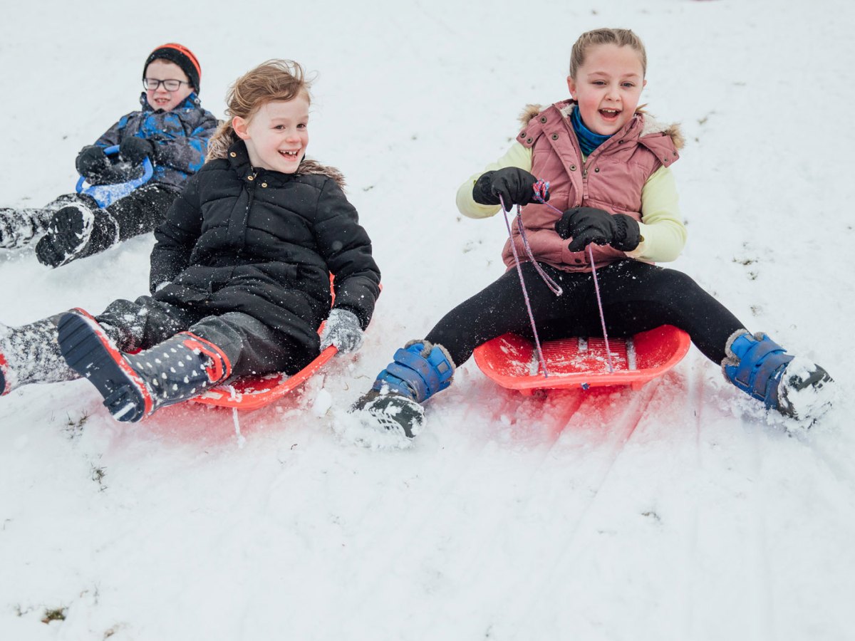 Drei Kinder rodeln einen verschneiten Berg runter.