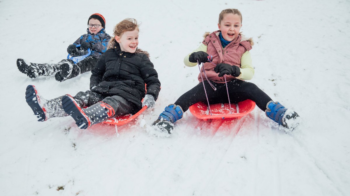 Drei Kinder rodeln einen verschneiten Berg runter.