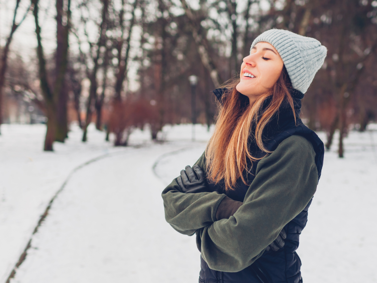 Junge Frau mit langen braunen Haaren und grauer Strickm&uuml;tze steht l&auml;chelnd in einem verschneiten Park und genie&szlig;t die Winterluft.