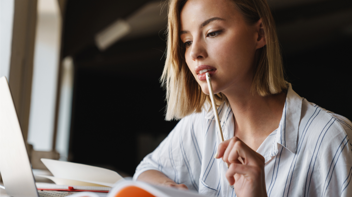 Frau an einem Schreibtisch, die nachdenklich einen Stift an den Mund hält und ein Buch liest.