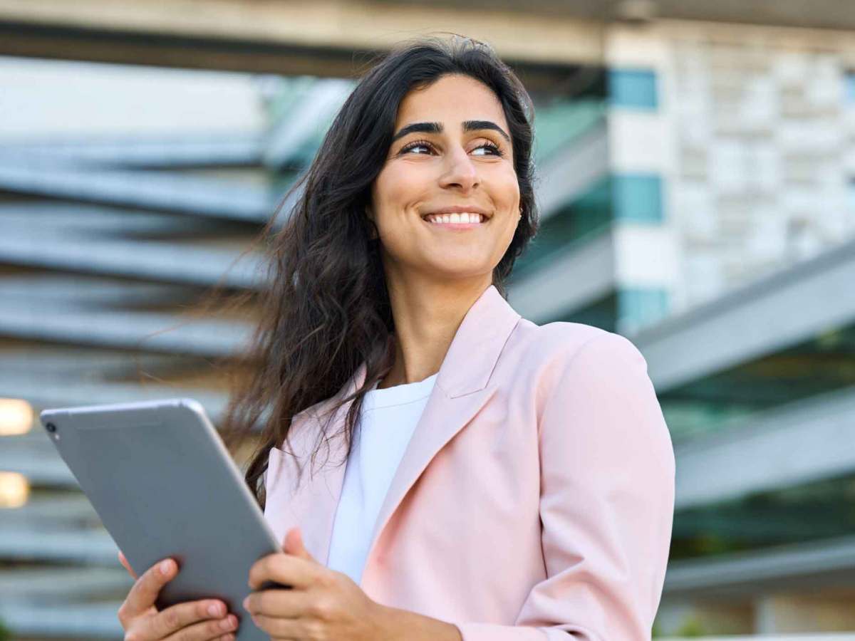 Frau mit rosa Blazer steht drau&szlig;en vor einem B&uuml;rogeb&auml;ude mit einem Tablet.