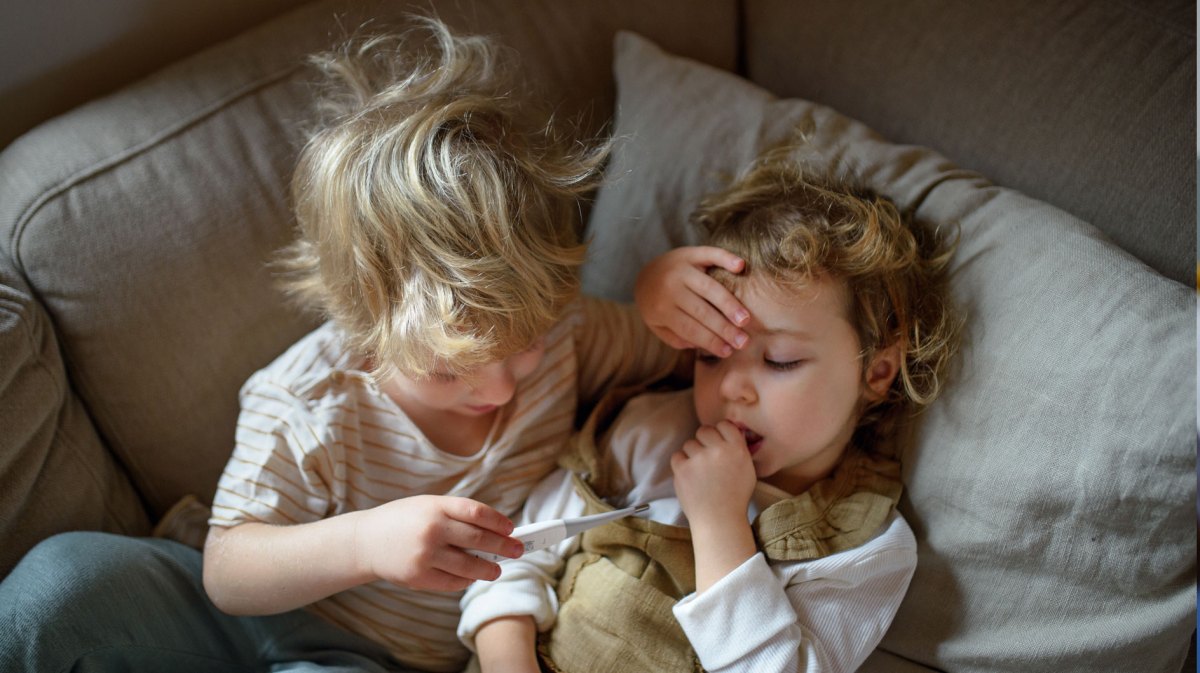 Zwei Kinder liegen im Bett. Der große Bruder, ca. 5 Jahre, legt seiner kleinen Schwester, ca. 3 Jahre, die Hand auf die Stirn und schaut auf das Fieberthermometer in seiner Hand.