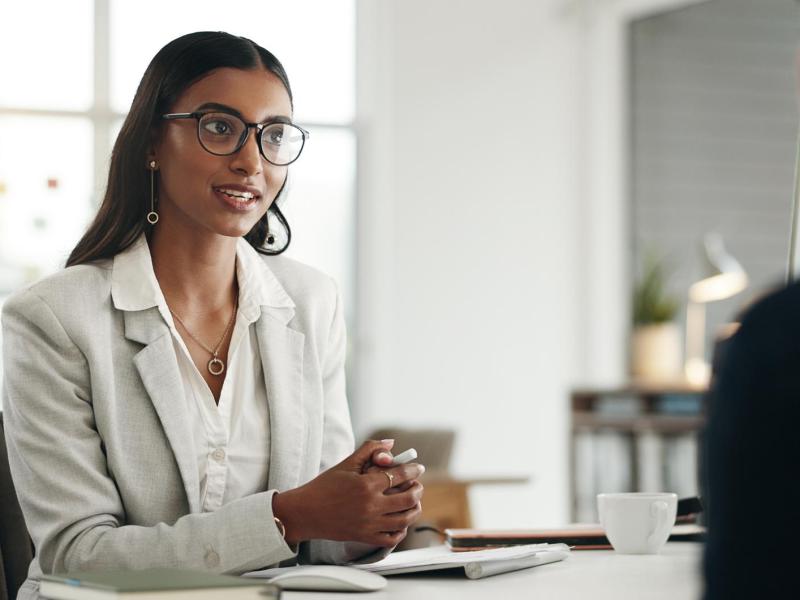 Frau mit dunklen Haaren, Brille und einem weißen Blazer sitzt bei einem Vorstellungsgespräch gegenüber von einem Personaler.