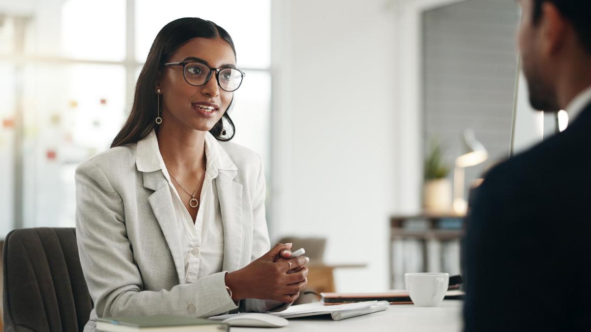 Frau mit dunklen Haaren, Brille und einem weißen Blazer sitzt bei einem Vorstellungsgespräch gegenüber von einem Personaler.