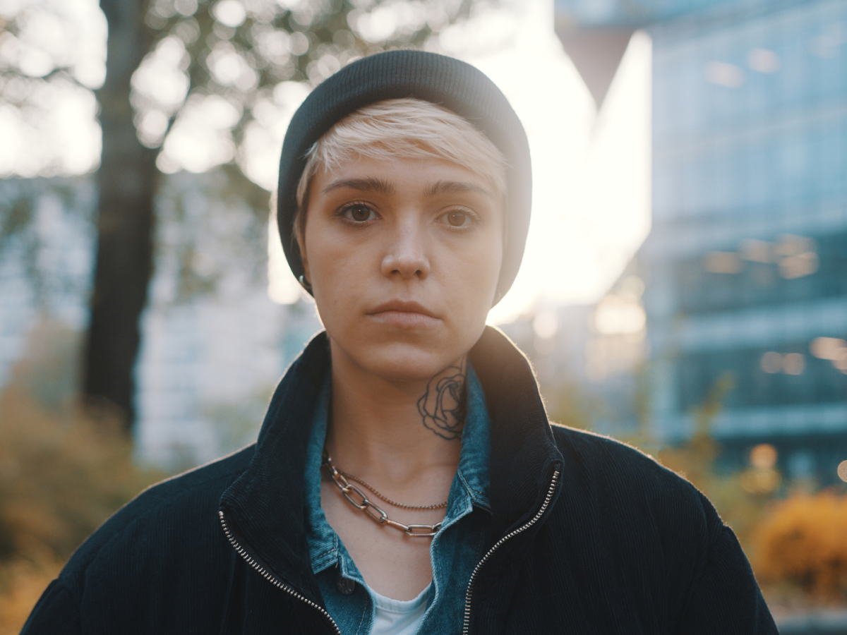 Junge Frau mit kurzem blondem Haar, schwarzer M&uuml;tze und t&auml;towiertem Hals blickt ernst in die Kamera, im Hintergrund herbstliche B&auml;ume und Glasfassaden.