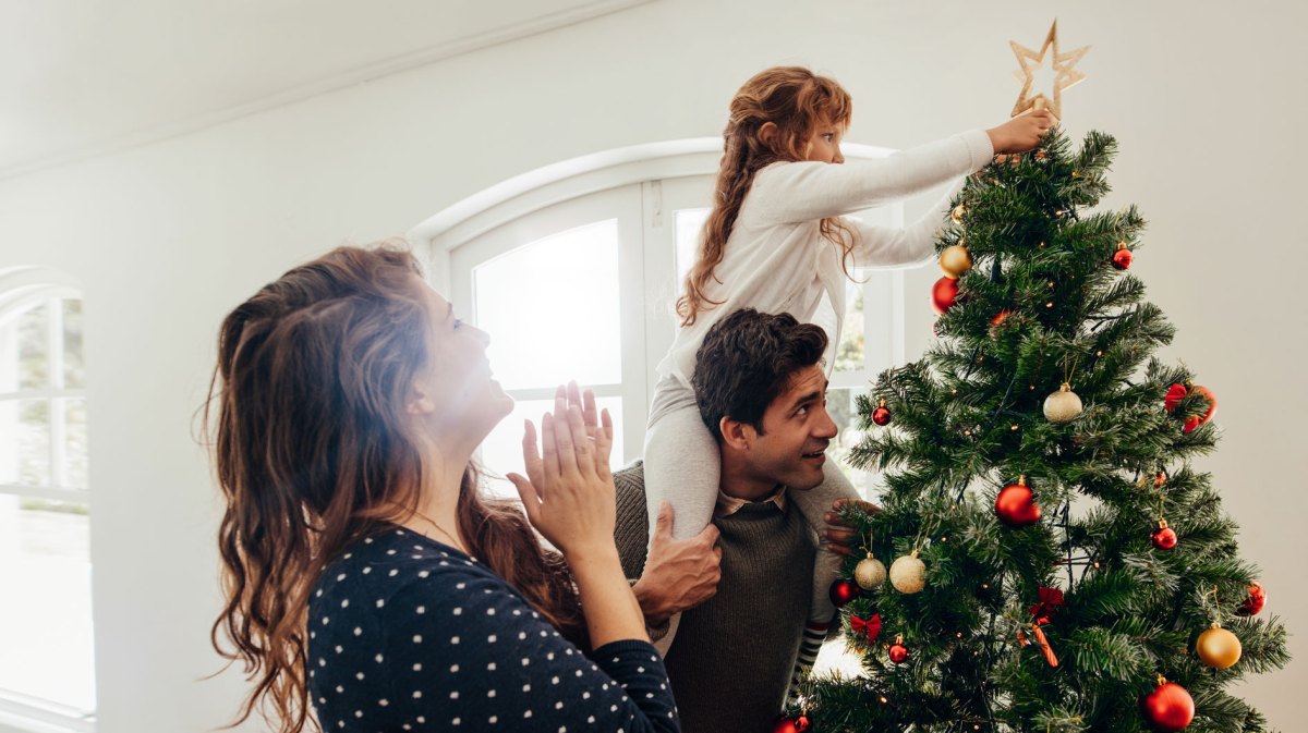 Mutter, Vater und Tochter haben den Weihnachtsbaum geschmückt. Die Tochter, auf den Schultern des Vaters sitzend, steckt dem Baum die Stern-Spitze an.