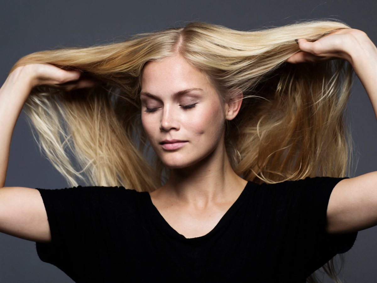Junge blonde Frau in schwarzem Shirt h&auml;lt ihr langes Haar mit geschlossenen Augen seitlich hoch vor dunkelgrauem Hintergrund.