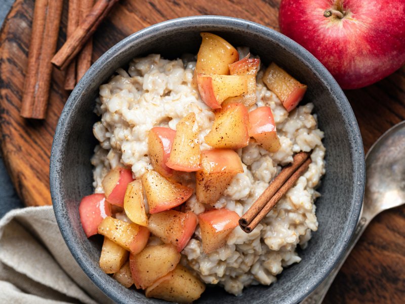 Apfel-Zimt-Porridge in einer schwarzen Schüssel auf einem Holzbrett. Ein Apfel, ein Löffel & Zimtstangen liegen daneben.