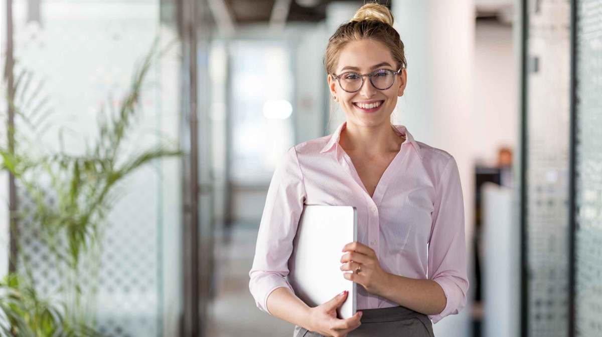 Frau mit Brille steht im Büro und hält ein Notebook unter dem Arm.