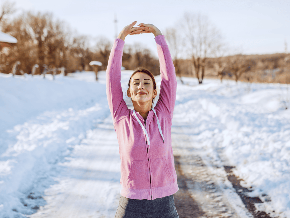 Eine junge Frau mit braunem Haar und rosa Kapuzenjacke streckt sich l&auml;chelnd auf einem verschneiten Weg in der Wintersonne.