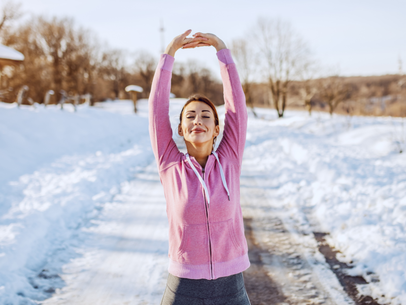 Eine junge Frau mit braunem Haar und rosa Kapuzenjacke streckt sich lächelnd auf einem verschneiten Weg in der Wintersonne.