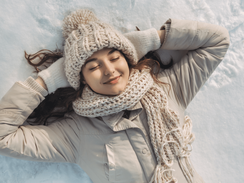 Glückliche Frau in beiger Winterjacke, Strickmütze und Schal liegt entspannt mit verschränkten Armen auf schneeweißem Boden.