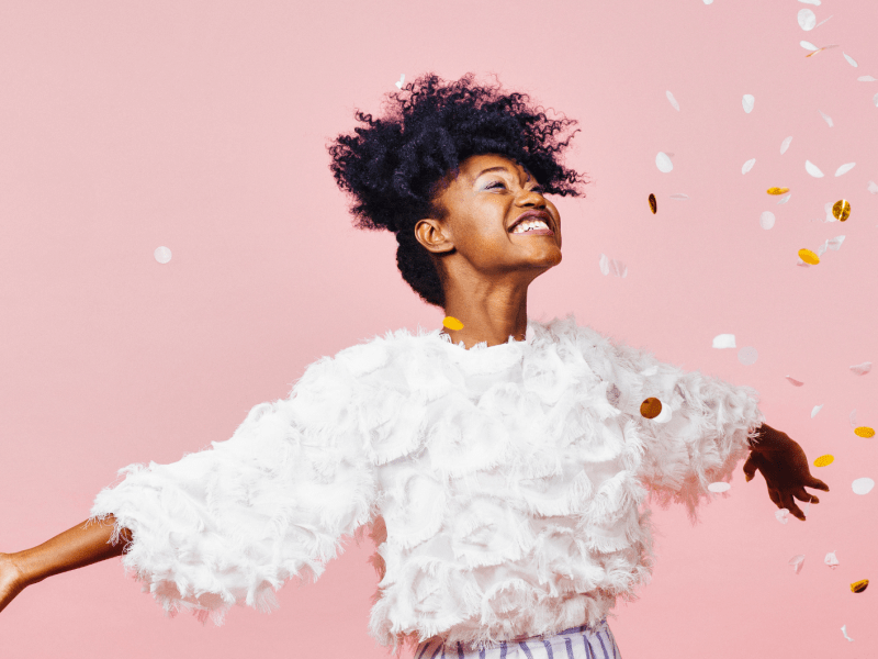 Junge Frau mit lockigem Afro und weißem Fransenshirt lacht strahlend mit ausgestreckten Armen, umgeben von Konfetti vor rosa Hintergrund.