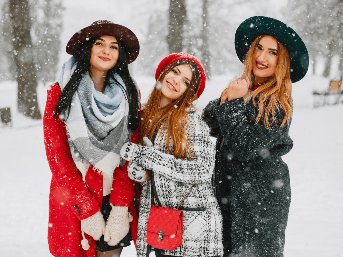 Drei Frauen im Schnee, mit Wintermantel und H&uuml;ten, die allesamt in die Kamera l&auml;cheln, w&auml;hrend es um sie herum schneit.