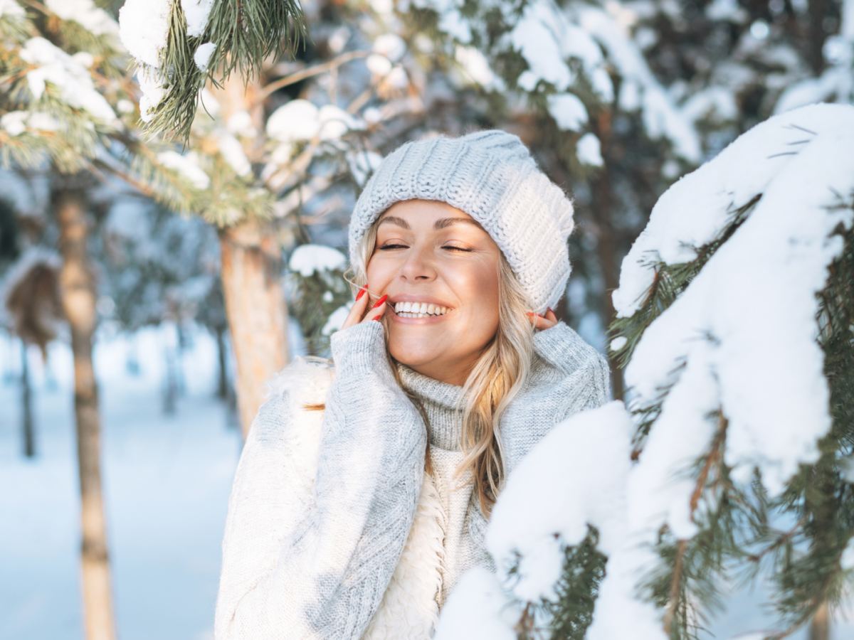 Junge Frau mit strahlendem L&auml;cheln und rotem Nagellack lehnt sich im warmen Winter-Outfit an einen schneebedeckten Baum.