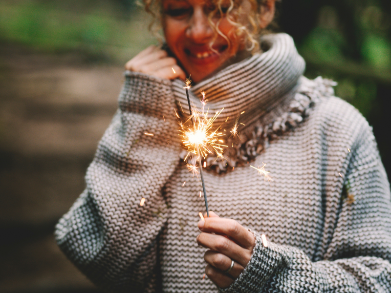 Eine lachende junge Frau mit lockigem blondem Haar hält eine brennende Wunderkerze, trägt einen groben Wollpullover im Wald.