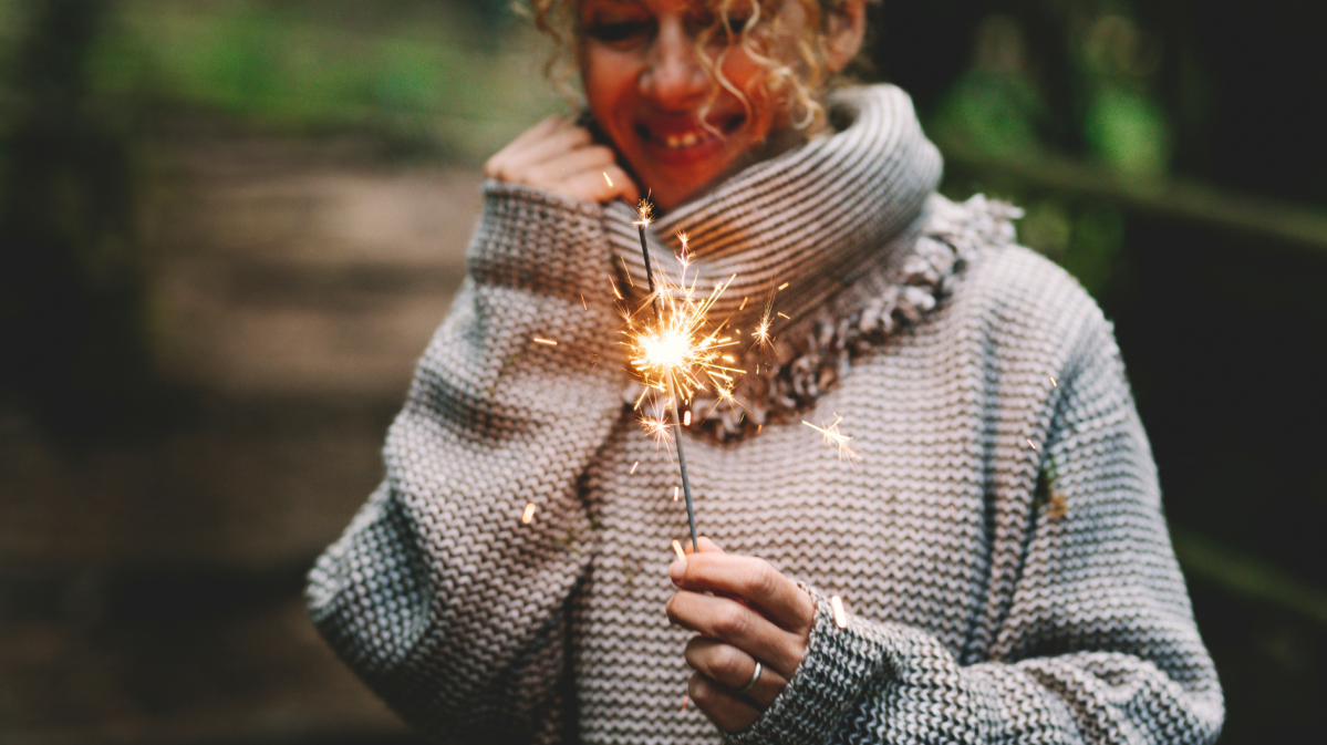 Eine lachende junge Frau mit lockigem blondem Haar hält eine brennende Wunderkerze, trägt einen groben Wollpullover im Wald.