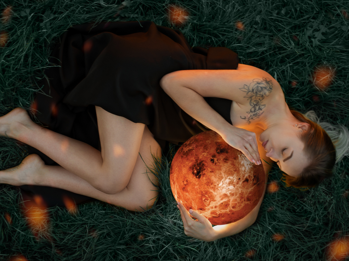 Eine junge Frau in schwarzem Kleid liegt barfu&szlig; im Gras, die Augen geschlossen, mit einem leuchtenden Planeten in ihren Armen.