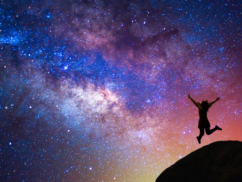 Person auf Hügel springt freudig in die Luft, Milchstraße und bunte Lichter am Himmel