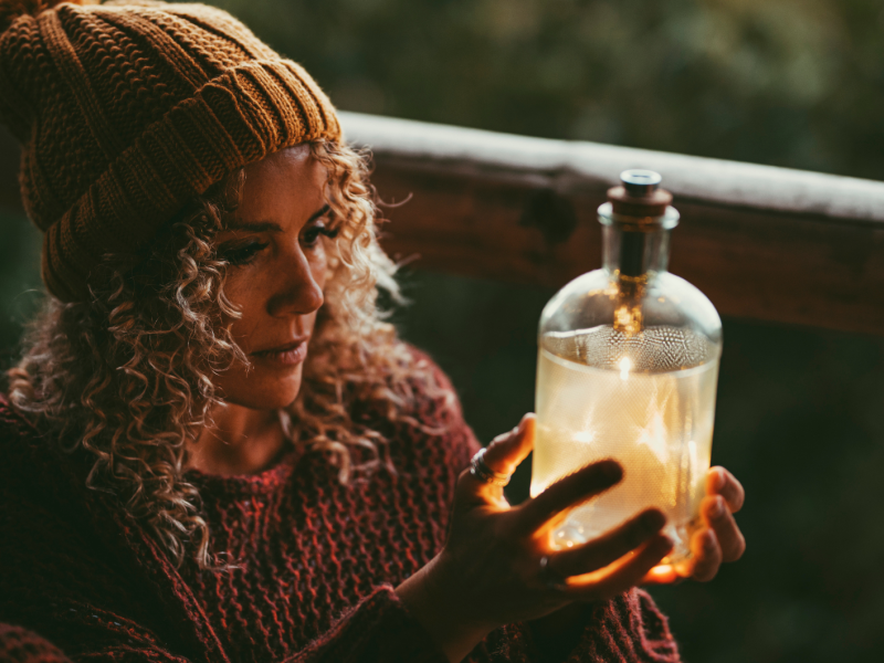 Eine junge Frau mit lockigem Haar und gelber Mütze betrachtet nachdenklich eine leuchtende Flasche in ihren Händen.