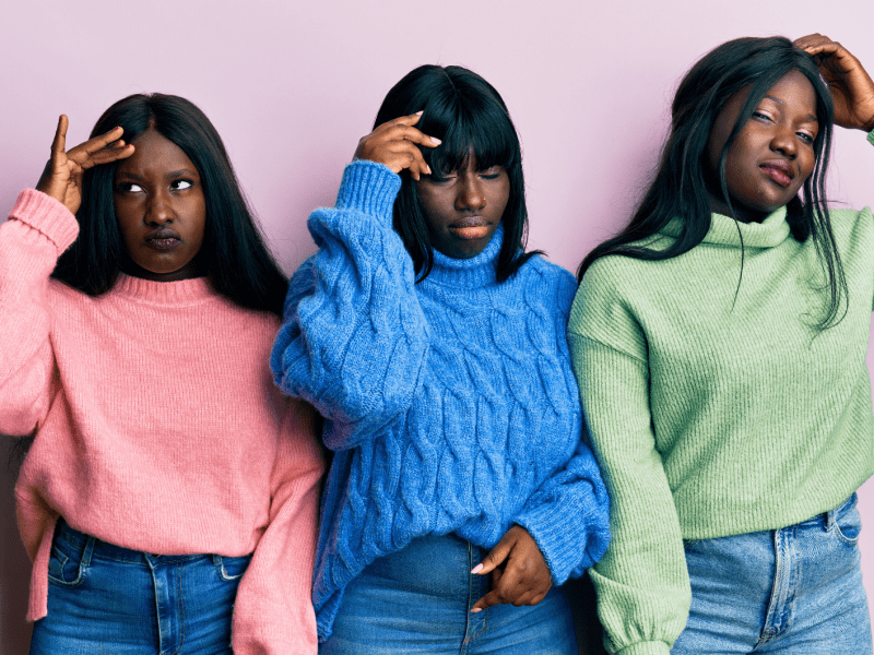 Drei junge Frauen mit langen schwarzen Haaren und ernsten Gesichtsausdrücken tragen bunte Strickpullover vor einer rosa Wand.