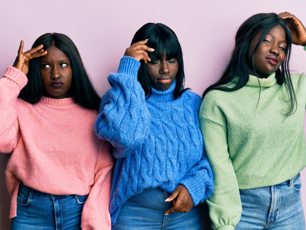 Drei junge Frauen mit langen schwarzen Haaren und ernsten Gesichtsausdr&uuml;cken tragen bunte Strickpullover vor einer rosa Wand.