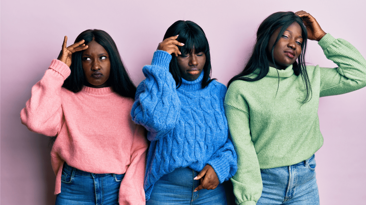 Drei junge Frauen mit langen schwarzen Haaren und ernsten Gesichtsausdrücken tragen bunte Strickpullover vor einer rosa Wand.